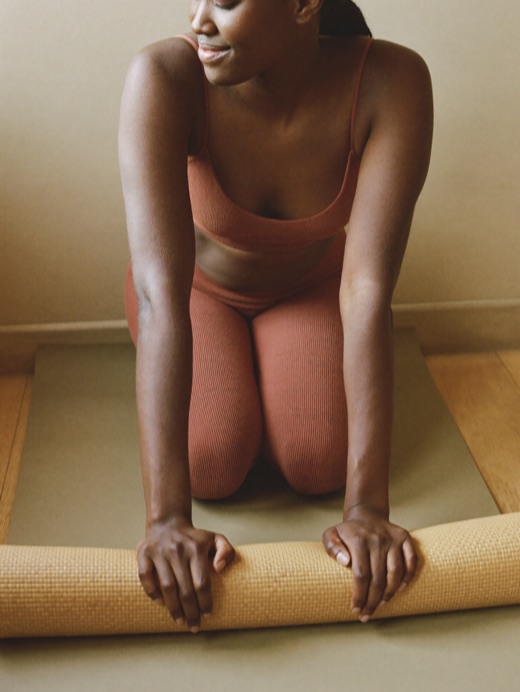 A woman kneels to roll up a woven yoga mat, wearing a clay bra and leggings.
