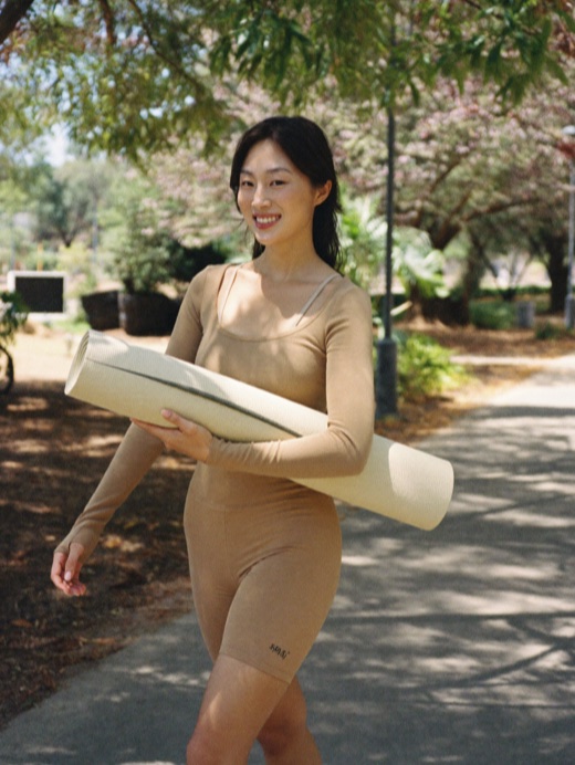 A woman walking through a park carrying a rolled yoga mat.