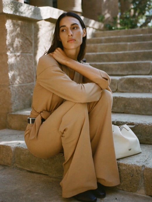 A woman sits on stone steps in dappled shade wearing an oat top and wide flare trousers.