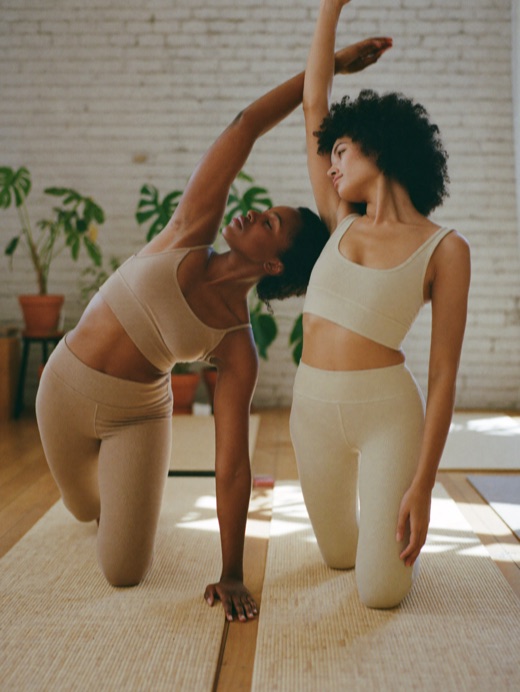 Two women stretch side by side on yoga mats in a sunlit studio.