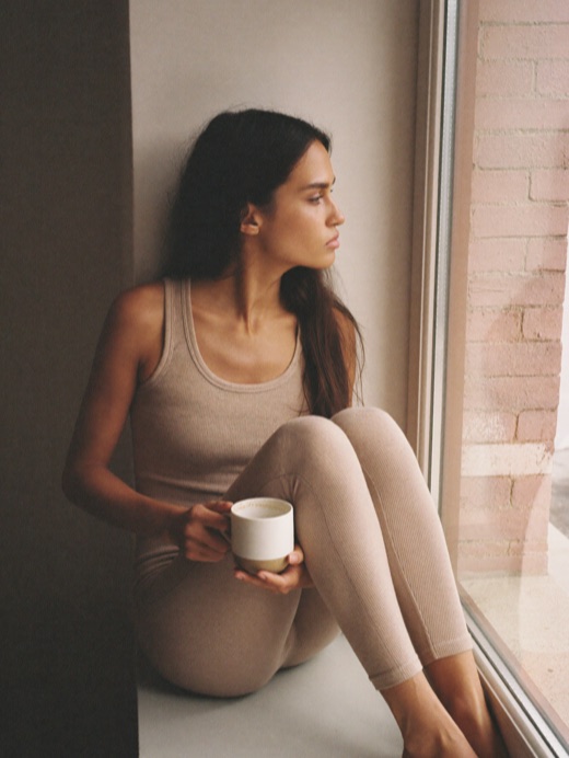 A woman sitting on a windowsill with a mug, in an oat tank and leggings.