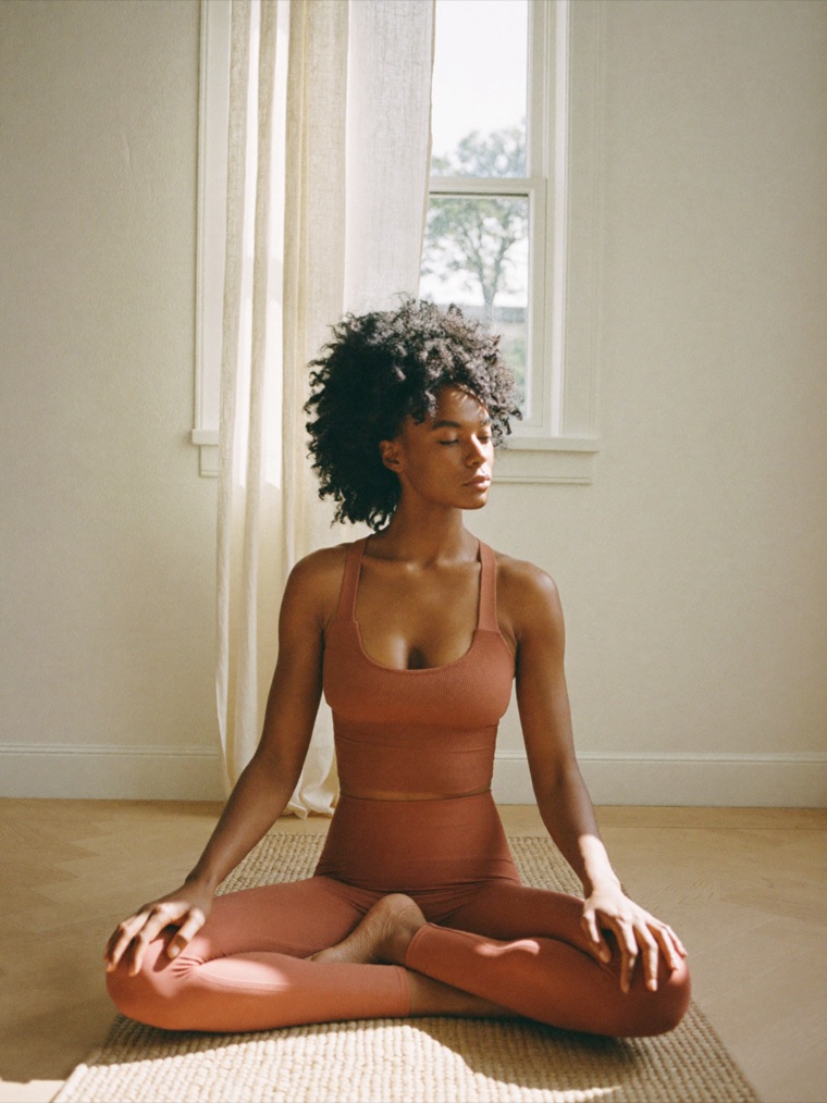 A woman sits cross-legged on a woven mat in a sunlit room, wearing a clay ribbed bra and matching leggings.